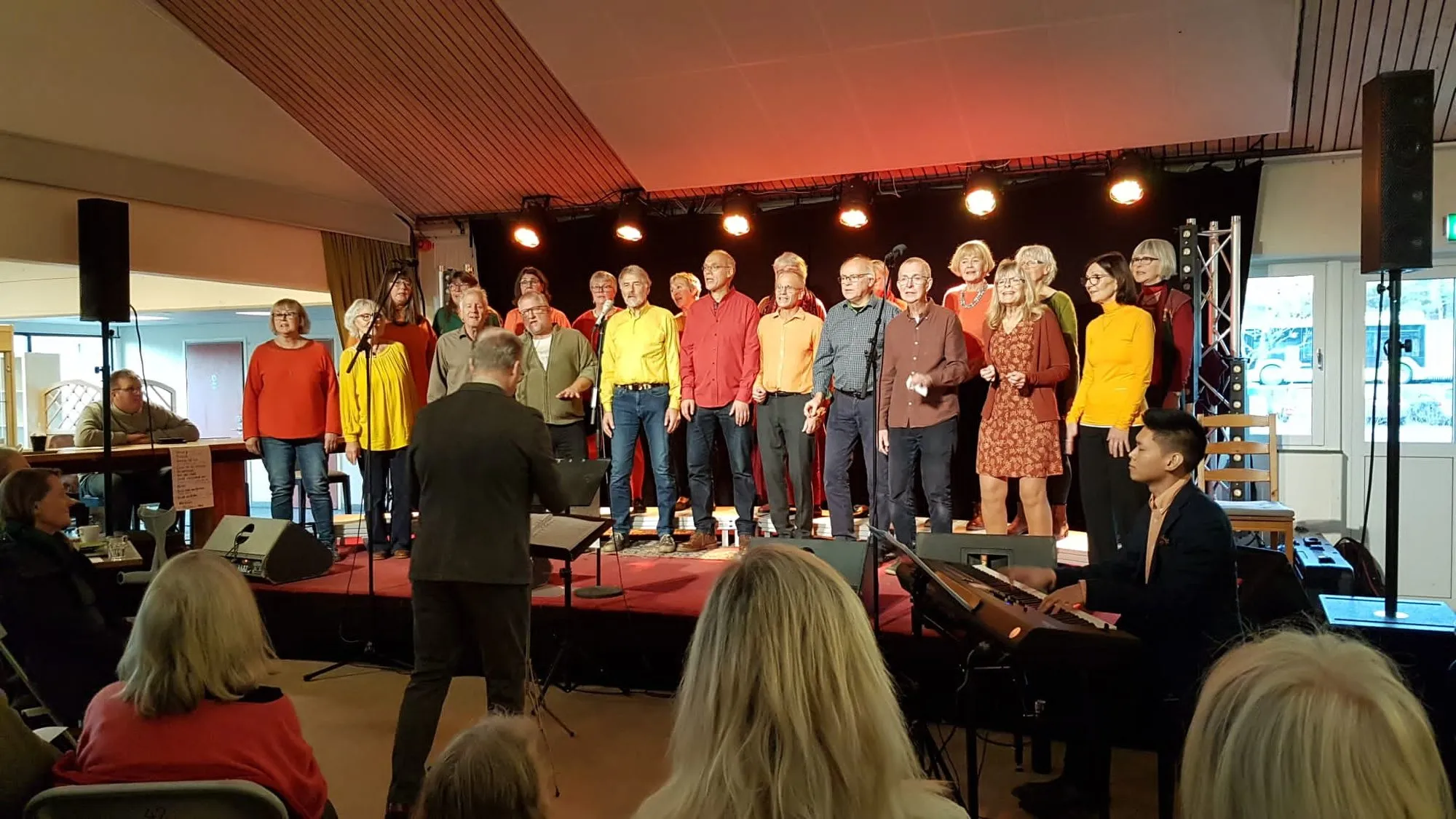 Kören i färgglada tröjor på scenen på Kulturoasen, med körledare och pianist framför.