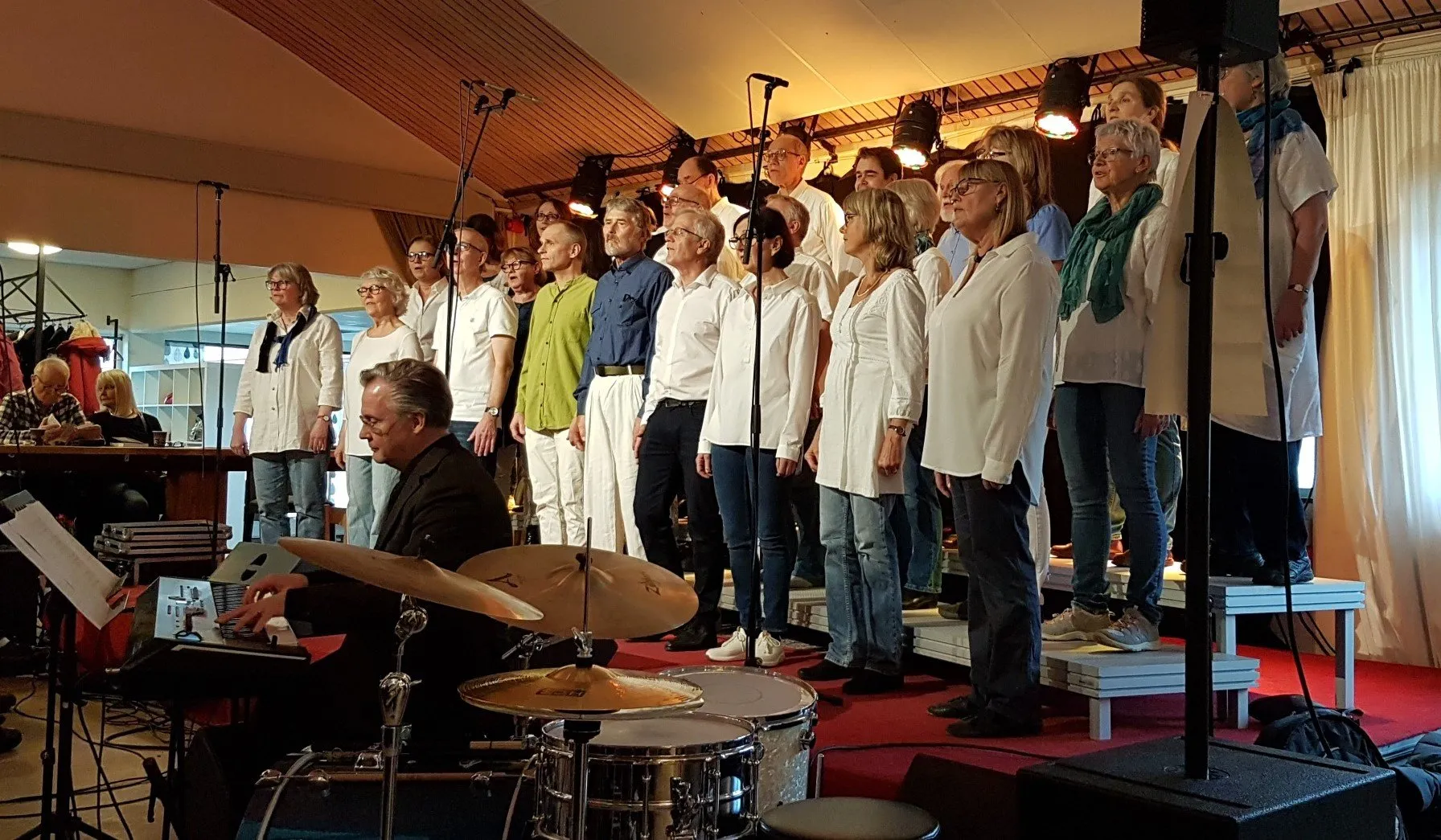 Kören i vita och gröna kläder på scenen, med musiker och trummor i förgrunden.